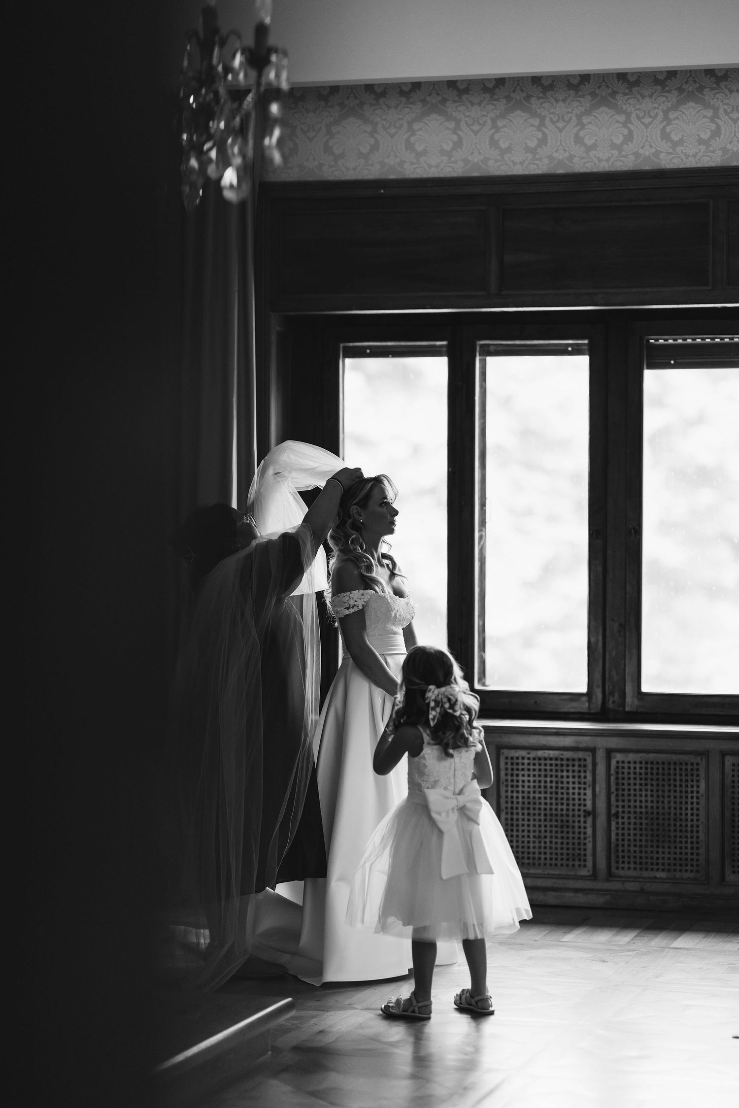 A touching black and white moment between figures in elegant attire by a window.