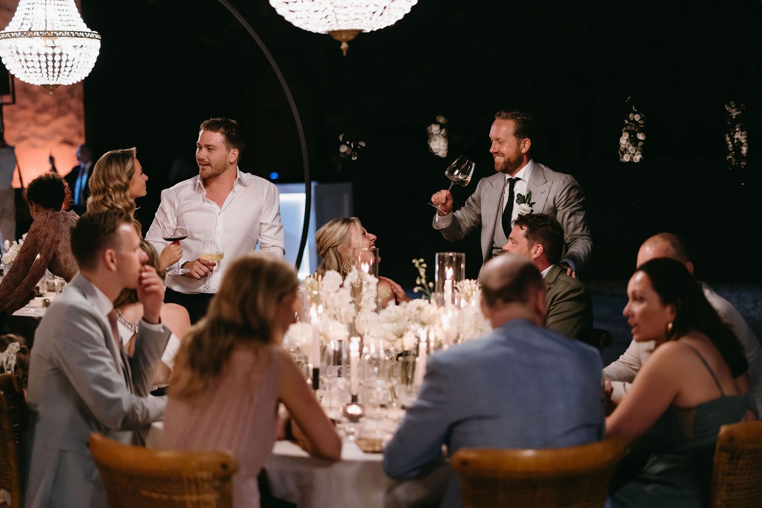 Evening wedding reception with guests gathered around candlelit tables under crystal chandeliers.