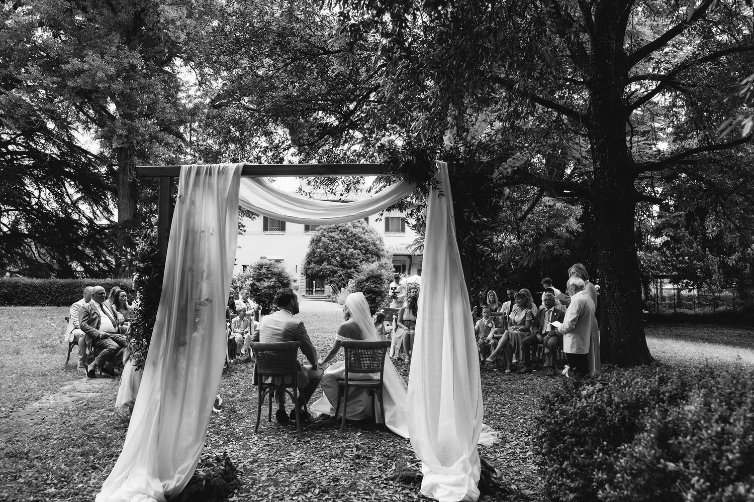Outdoor wedding ceremony setup with white drapes flowing between wooden posts in a garden setting.