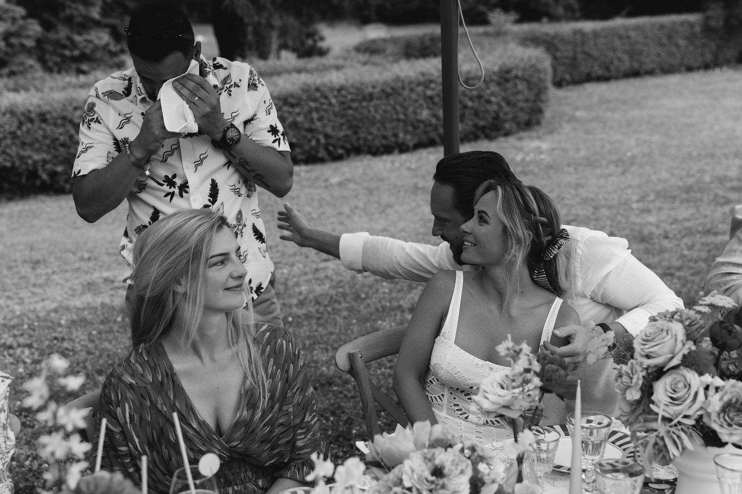 Guests enjoy a festive outdoor table setting with floral arrangements and laughter at a wedding celebration.
