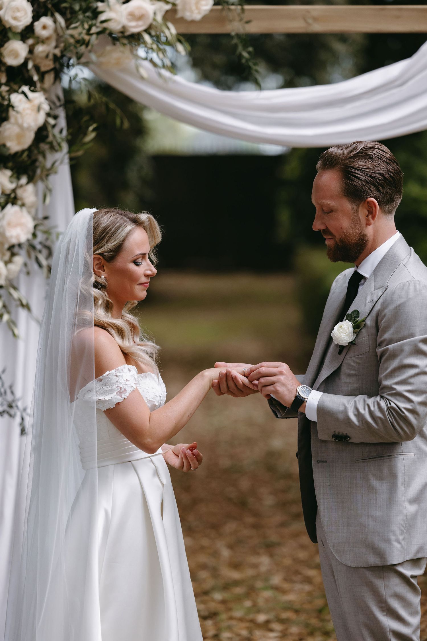 Couple exchanging rings under a white floral arch during an outdoor ceremony.