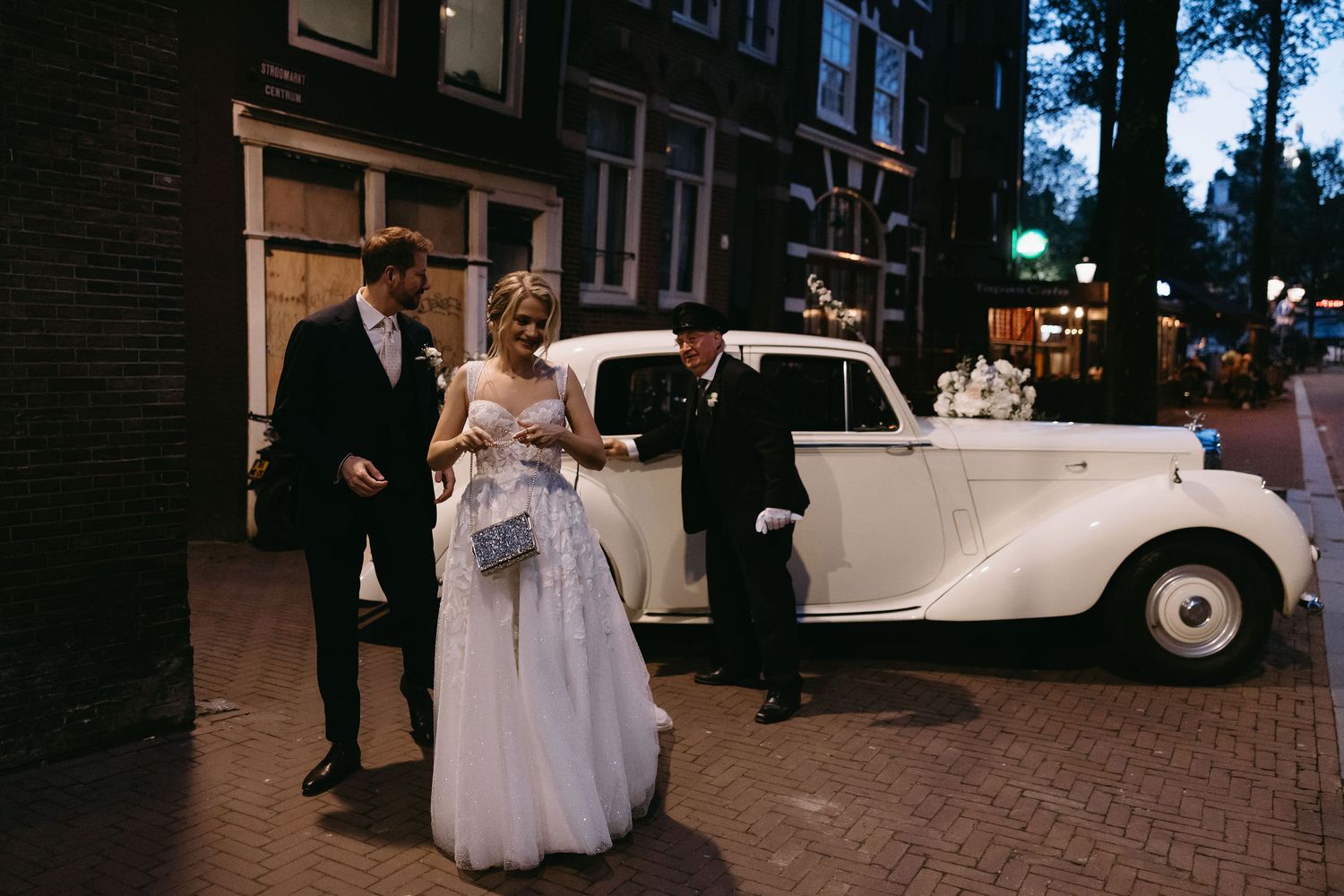 Wedding party gathers near classic white vintage car on city street at dusk.