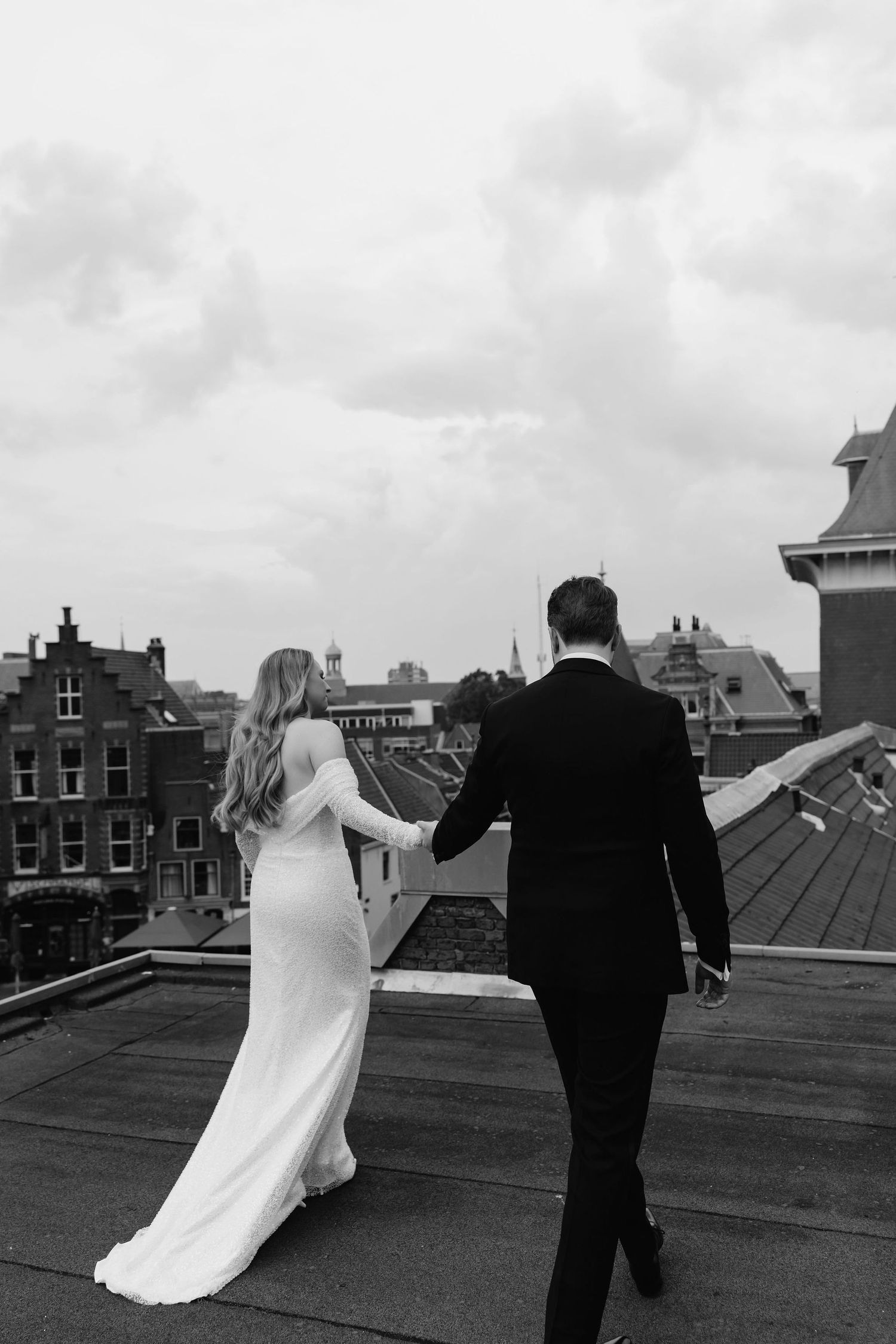 Black and white photo of couple walking on rooftop with cityscape in background.