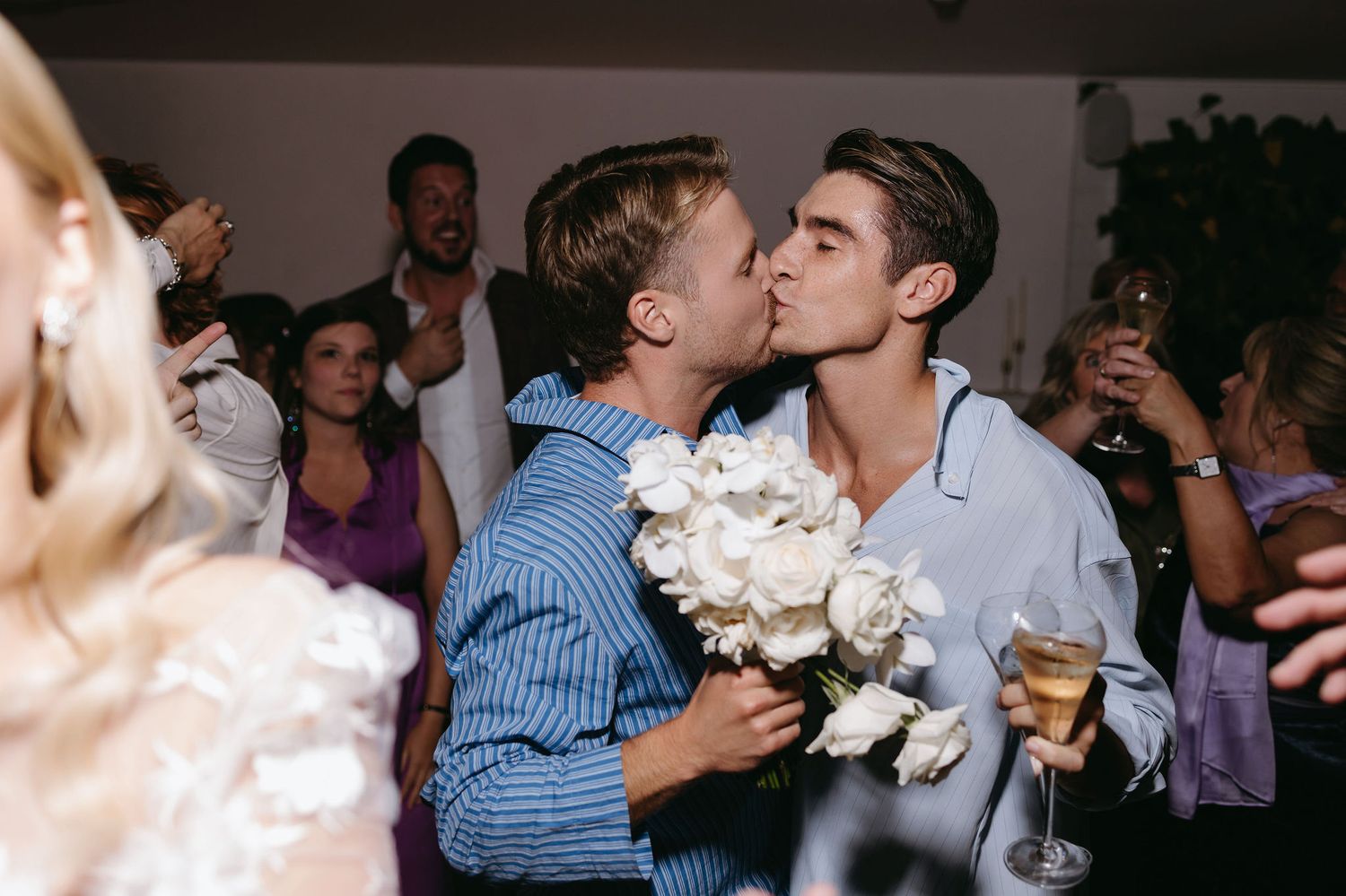 Two people share an affectionate moment at a celebration while holding a bouquet of white flowers.