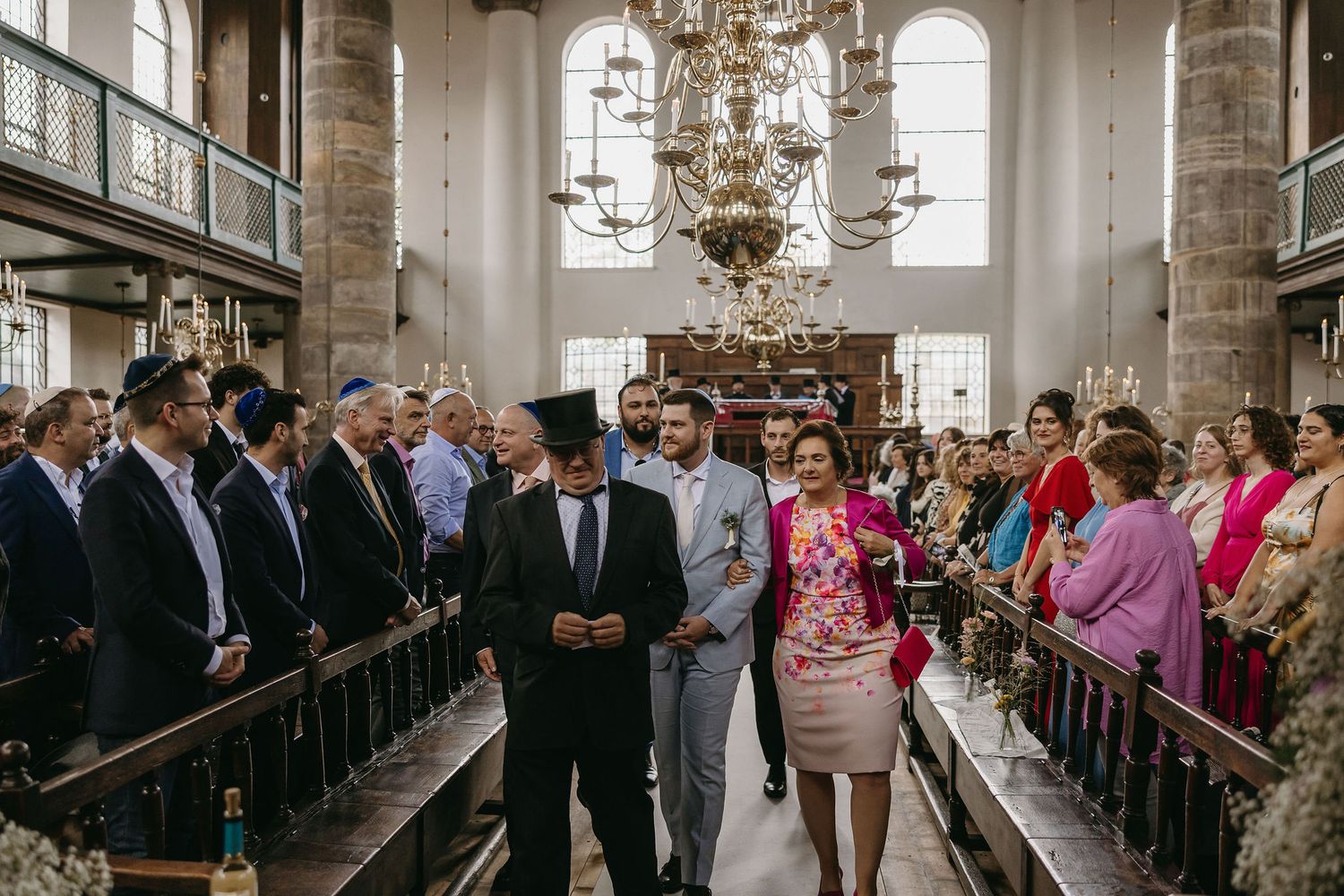 Wedding guests stand in church pews during a ceremony under an ornate chandelier.