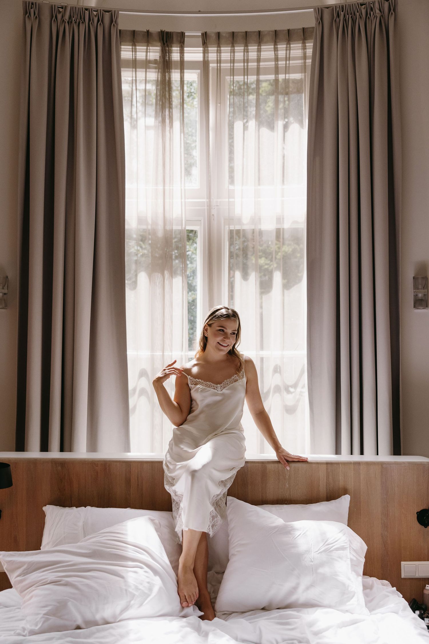Dreamy morning portrait of person in white slip dress sitting on bed by window with flowing curtains.