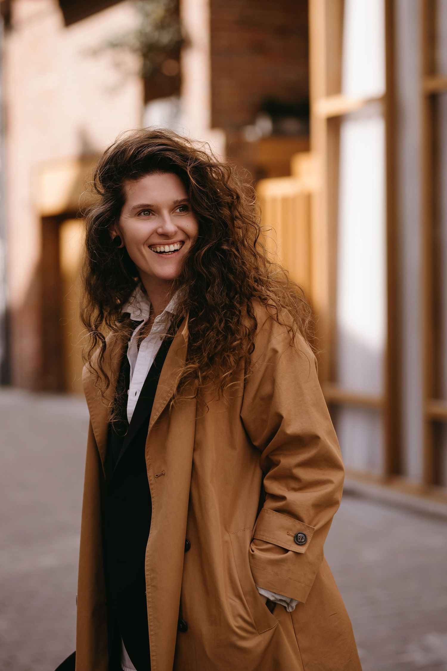 Person in tan trench coat smiling on urban street in warm sunlight.