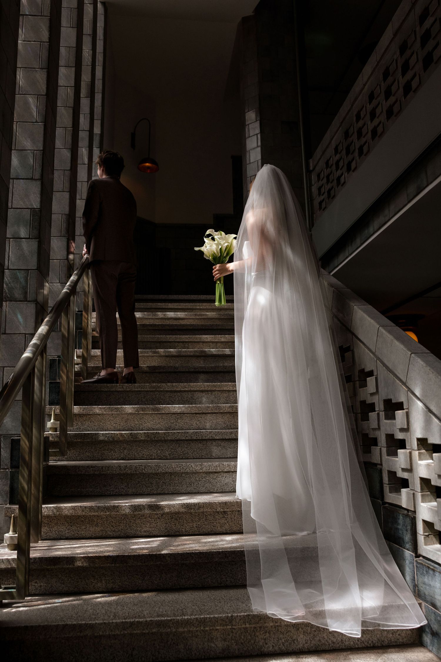 A bride in a flowing white veil holds a bouquet while ascending granite stairs behind a suited figure in dramatic lighting.