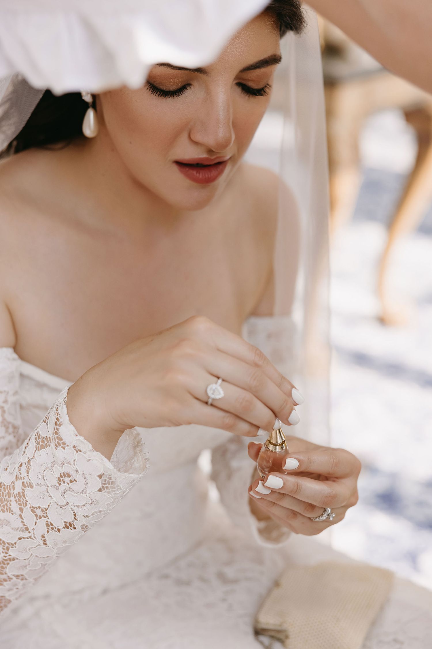 A bride in white lace dress holds a small perfume bottle while wearing an elegant diamond engagement ring.