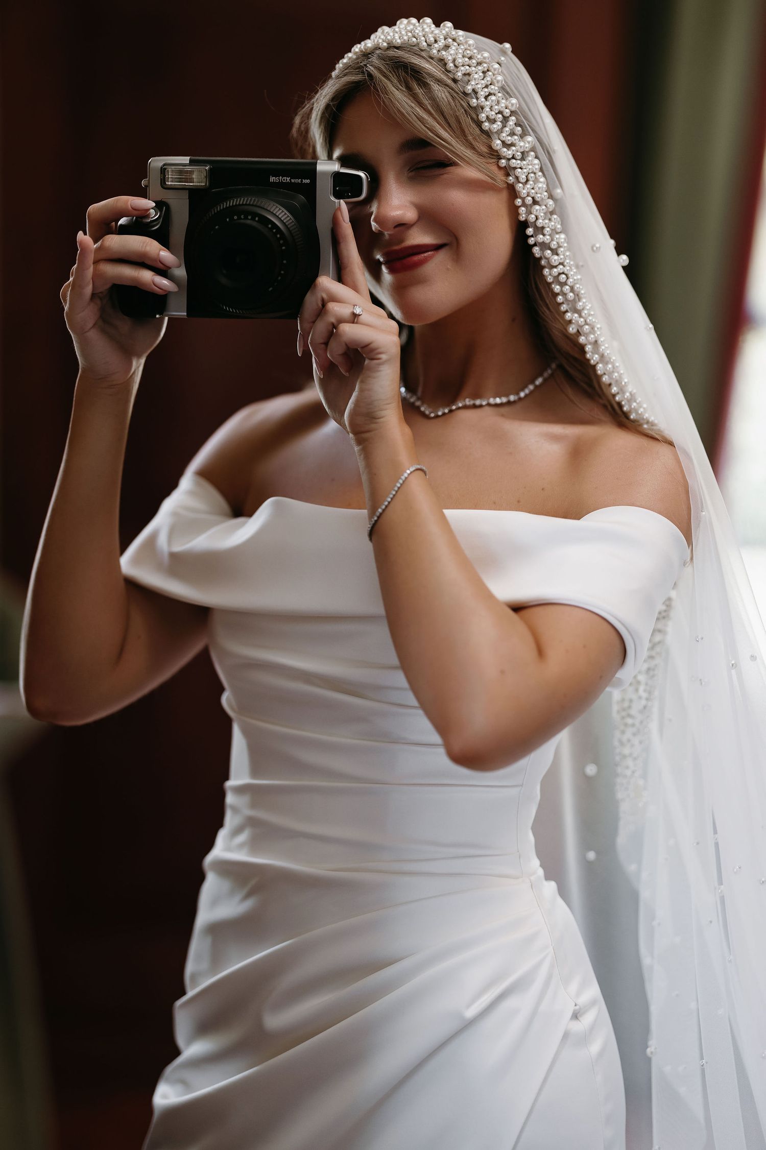 Bride in off-shoulder white wedding gown holds up a vintage film camera while wearing a long lace veil.