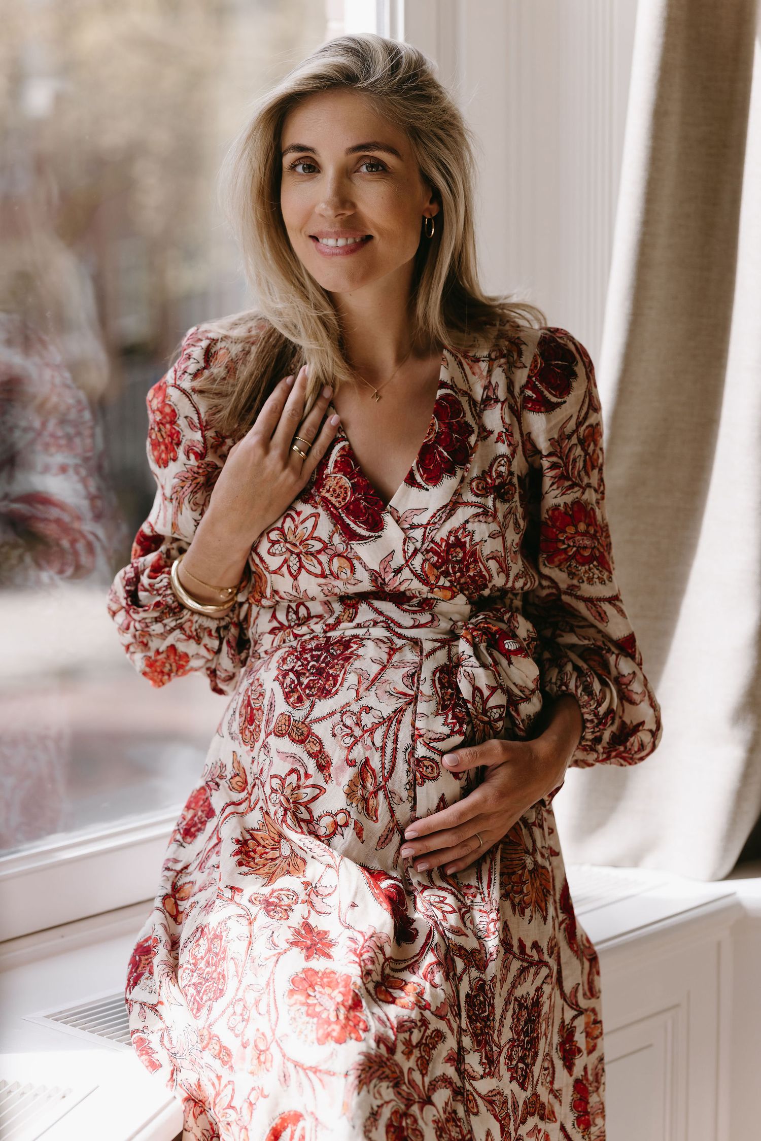 Woman in a red floral dress poses by bright windows in an indoor photoshoot.