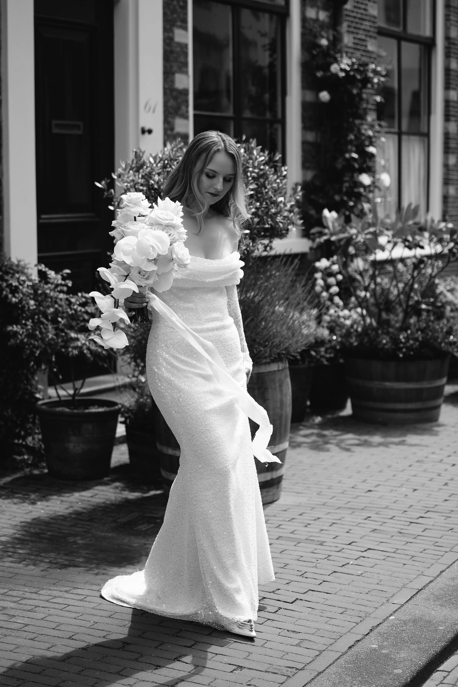 Black and white portrait of bride in fitted gown holding cascading white bouquet on brick pathway.