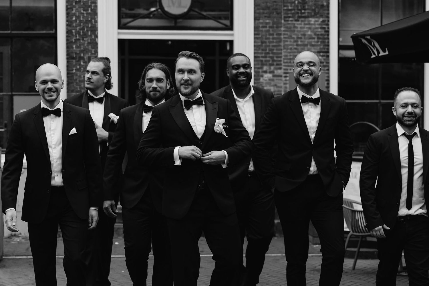 Group of groomsmen in black tuxedos and bowties posing together outside.