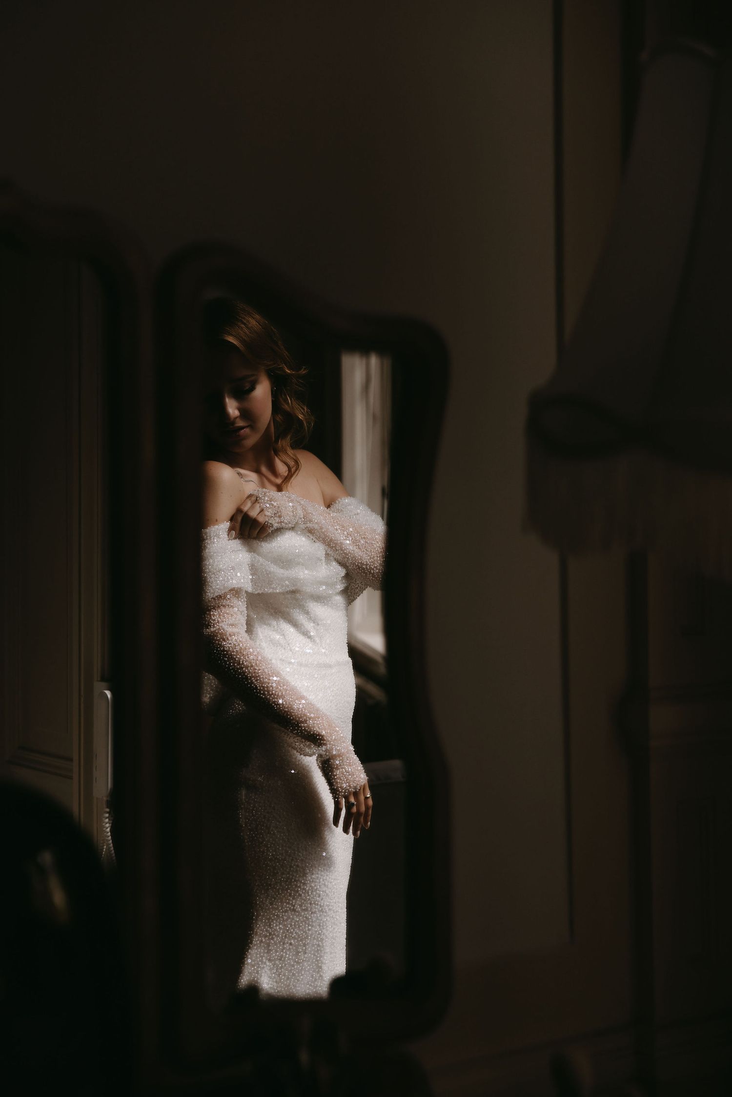 Dramatic window light illuminates wedding dress in moody bridal portrait.