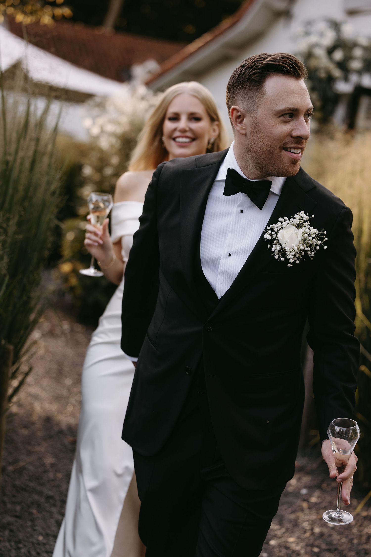 A wedding couple walking outdoors holding champagne glasses, with the bride in white and groom in a black tuxedo.
