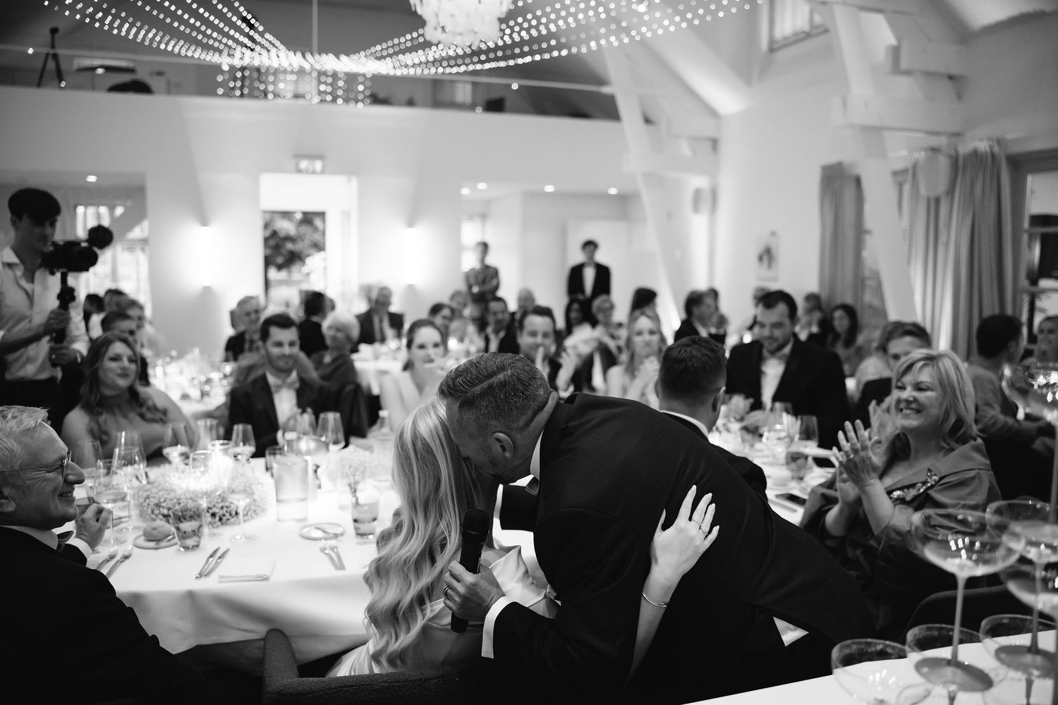 Black and white photo of wedding guests enjoying dinner at reception tables under string lights.