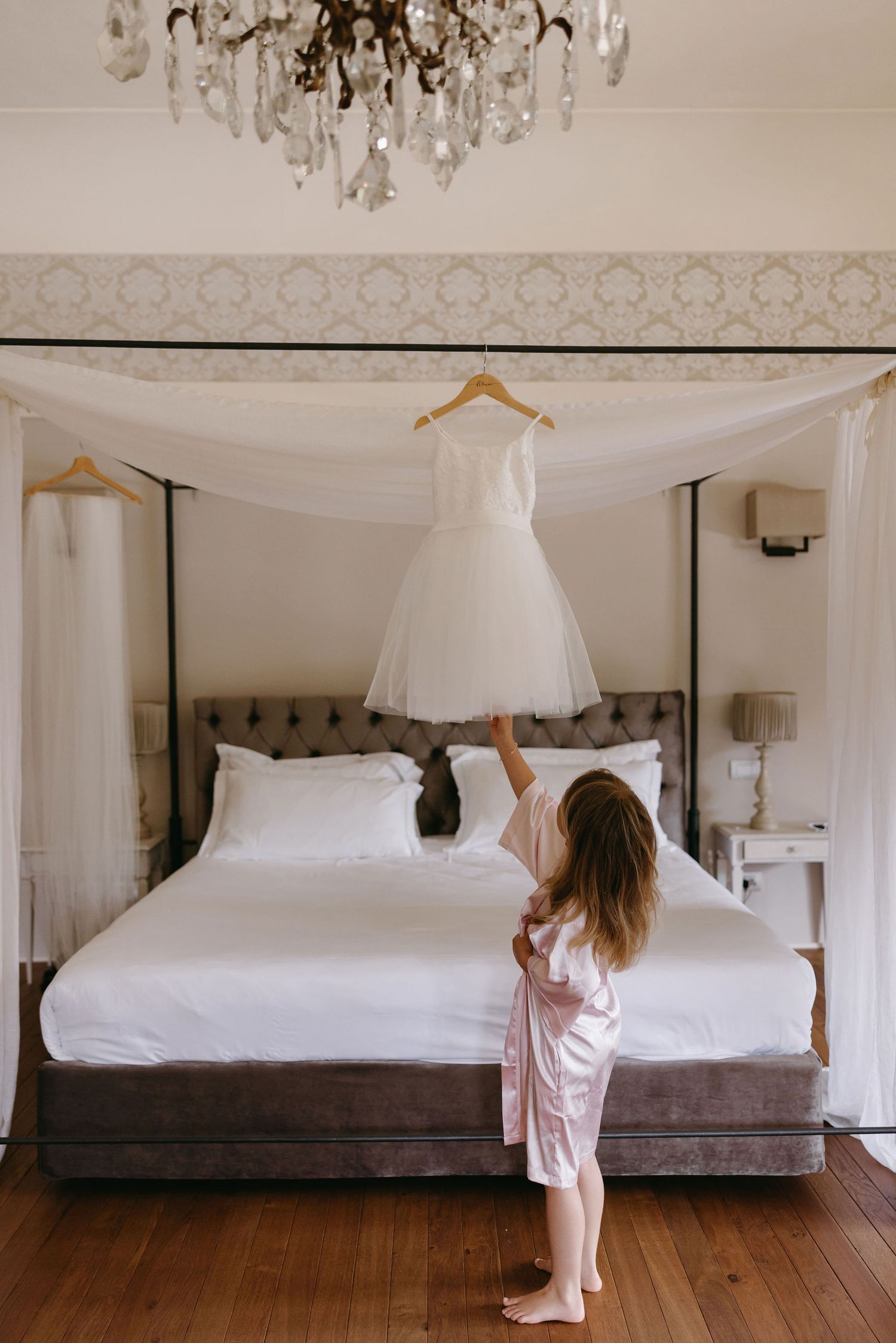 Short white wedding dress hanging from canopy bed beneath crystal chandelier.