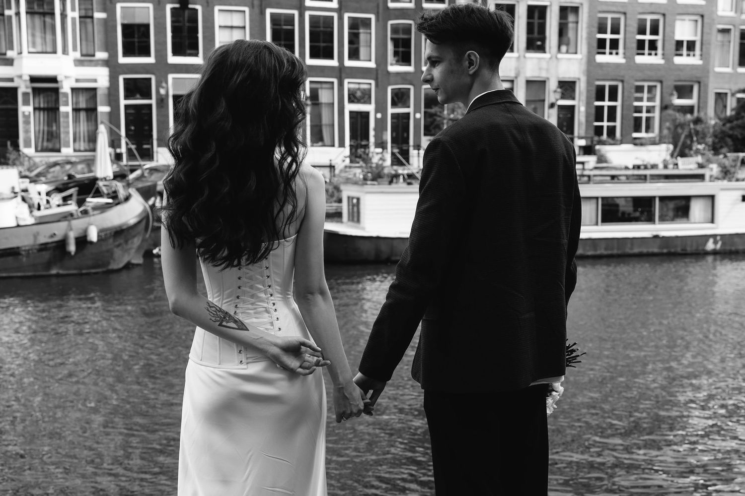 A couple in formal attire holds hands while standing along a canal in Amsterdam with classic buildings in background.