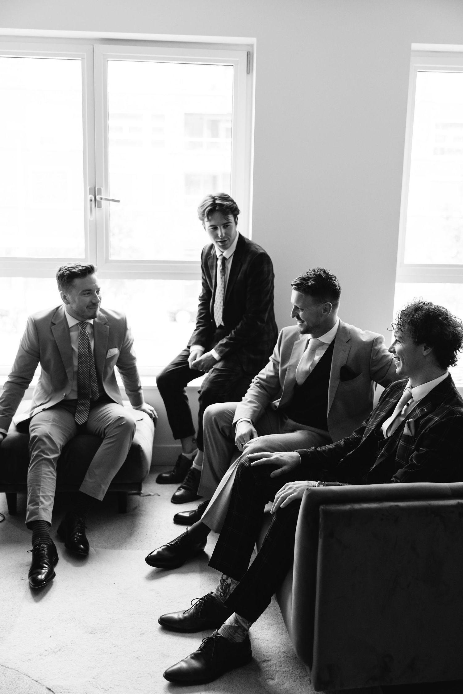 Groomsmen in formal suits sit casually by bright windows in modern room.