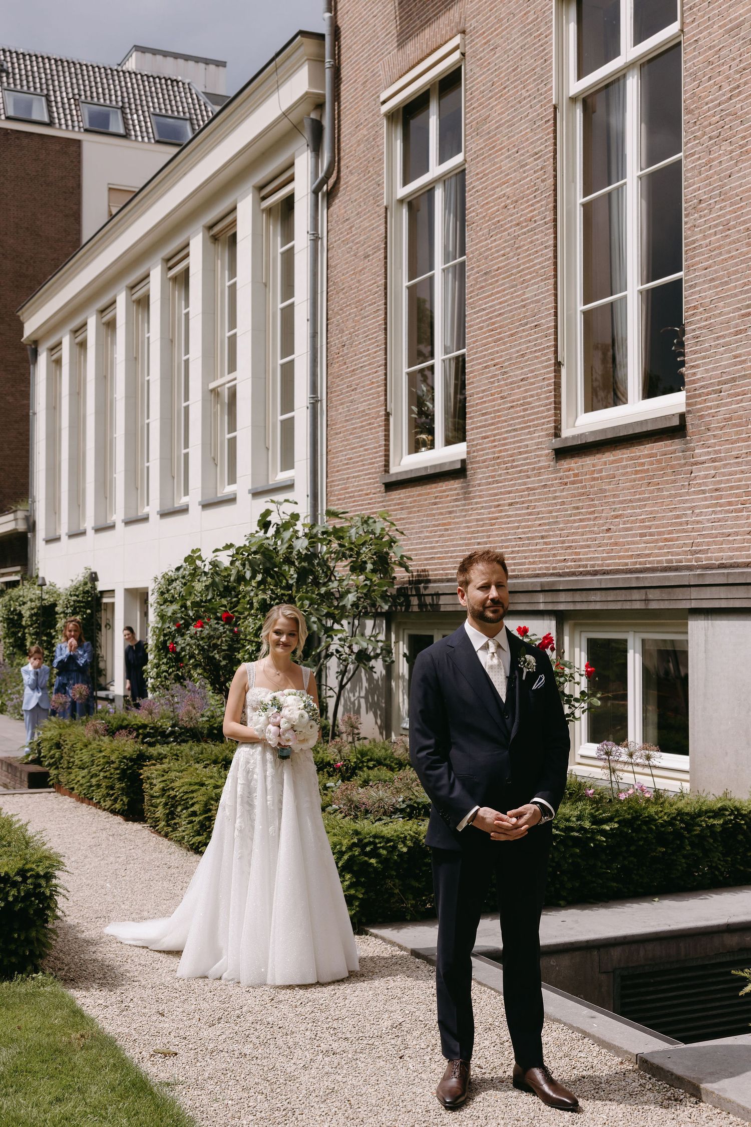 Wedding couple stands separately during first look moment outside brick building with garden path.