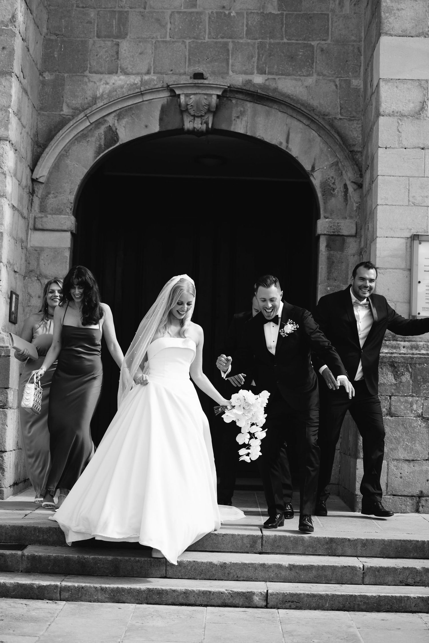 Wedding party exits stone church after ceremony through an arched doorway.