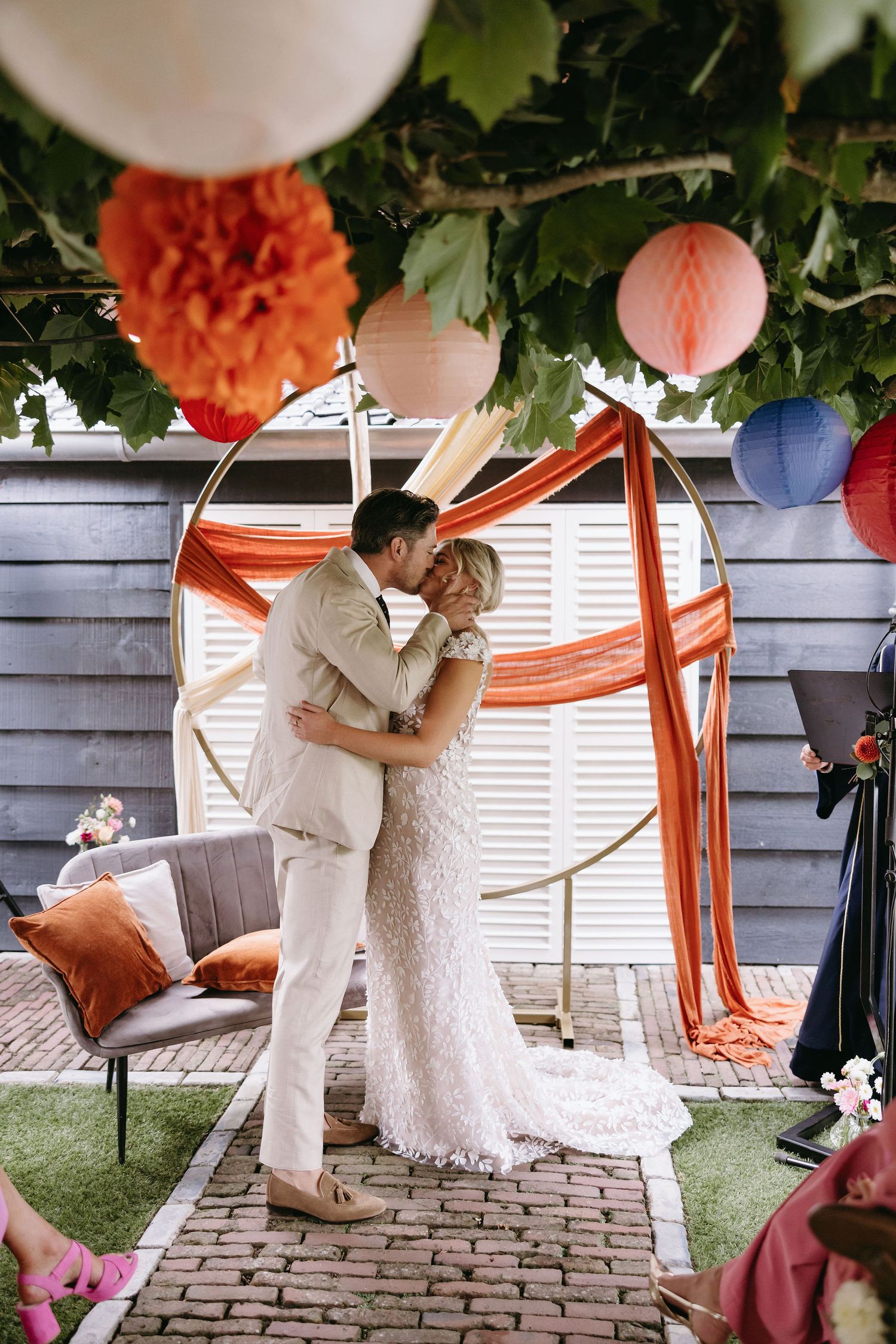 Newlyweds share intimate moment under coral and pink lanterns at backyard wedding ceremony.