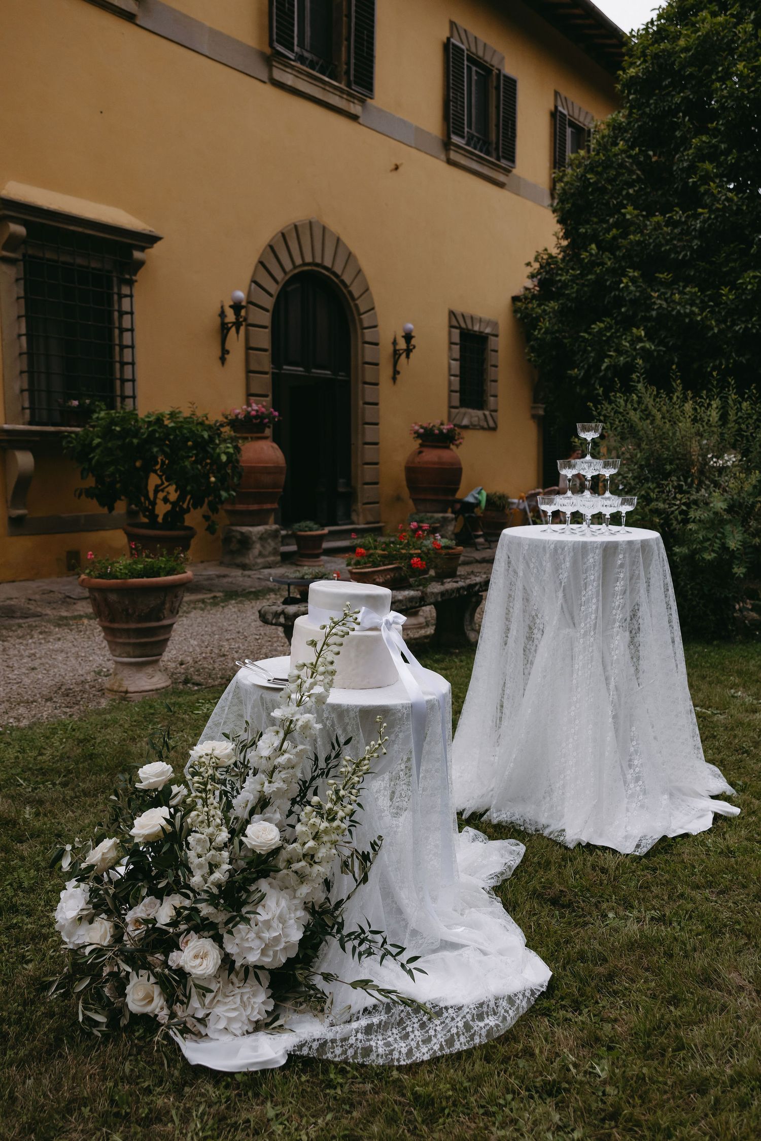 Elegant wedding cake display with white linens and floral arrangements outside a rustic Italian villa.