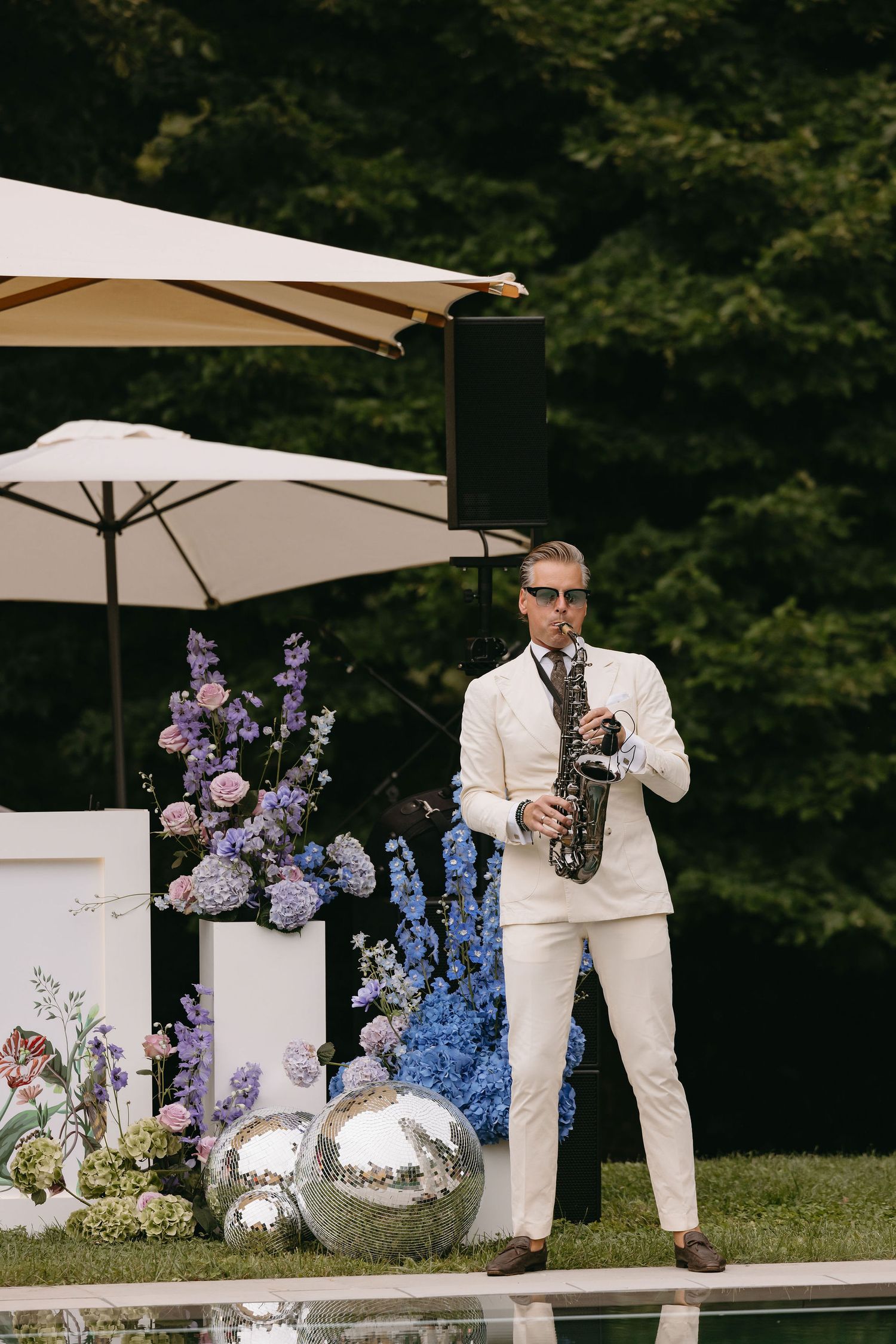 A saxophonist in a white suit performs outdoors next to purple floral arrangements and silver orbs.