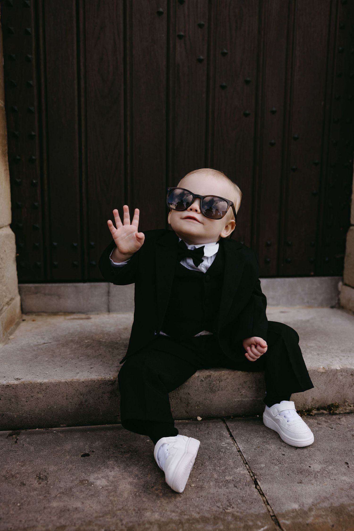 Playful wedding party member in formal attire sitting against dark wooden wall.