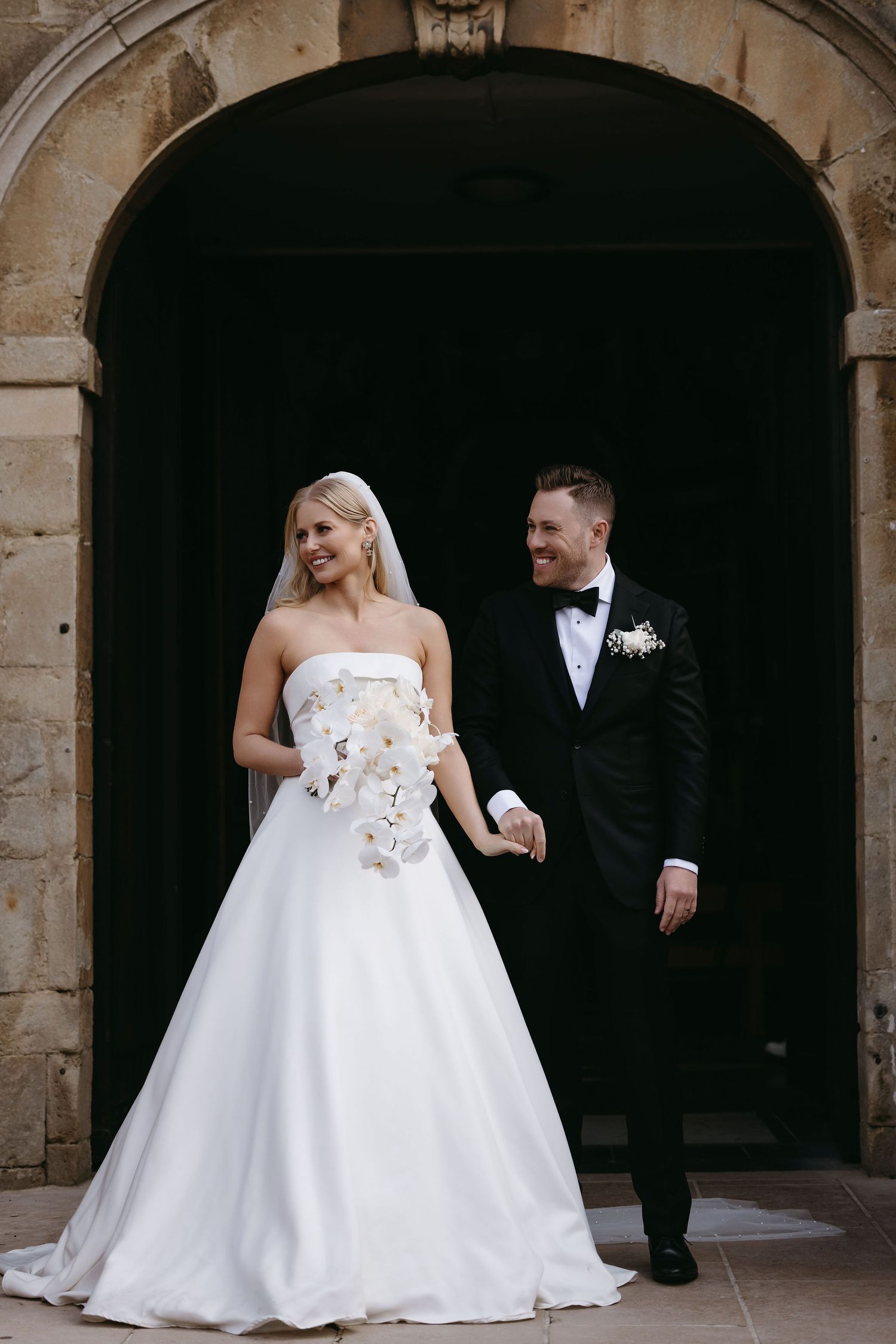 Couple in wedding attire pose under stone archway during formal portraits.