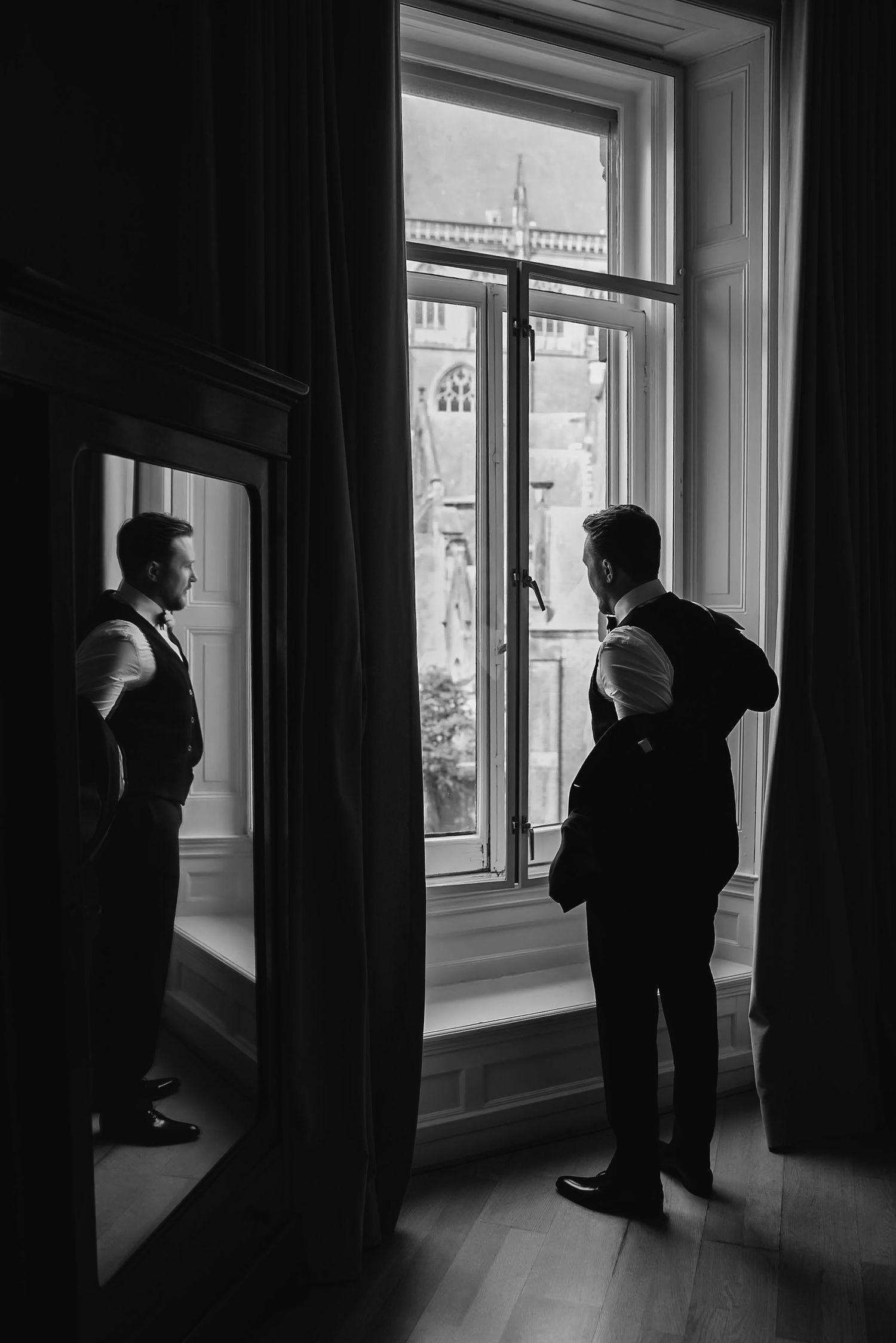 Two silhouetted figures stand by a tall window in a dramatic black and white composition.