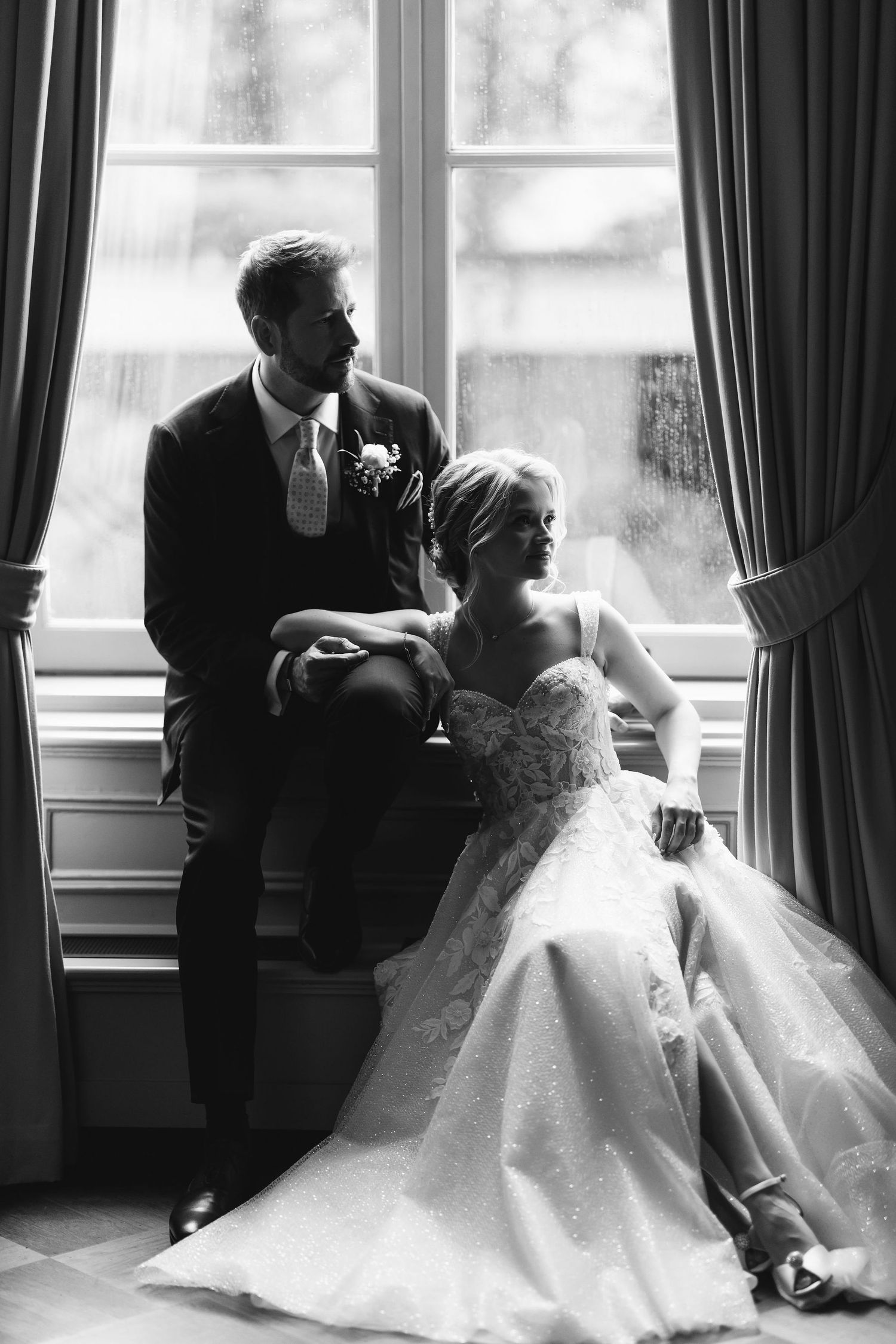 Artistic black and white portrait of newlyweds posing by a window in formal attire.