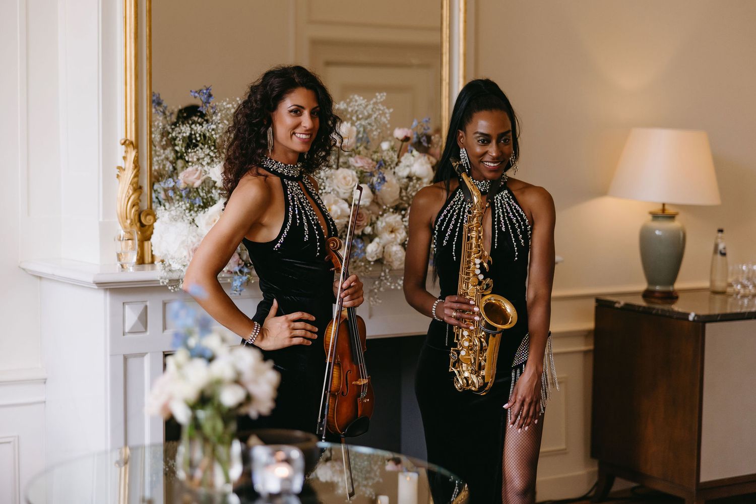 Two musicians pose elegantly with violin and saxophone in formal black dresses against a bright interior setting.