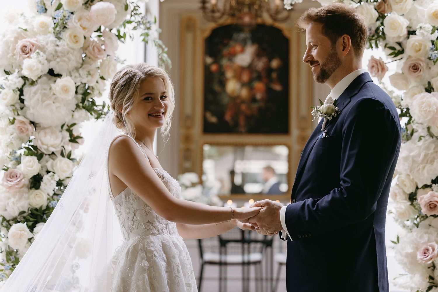 Wedding ceremony under an elegant white floral arch in an ornate indoor setting.