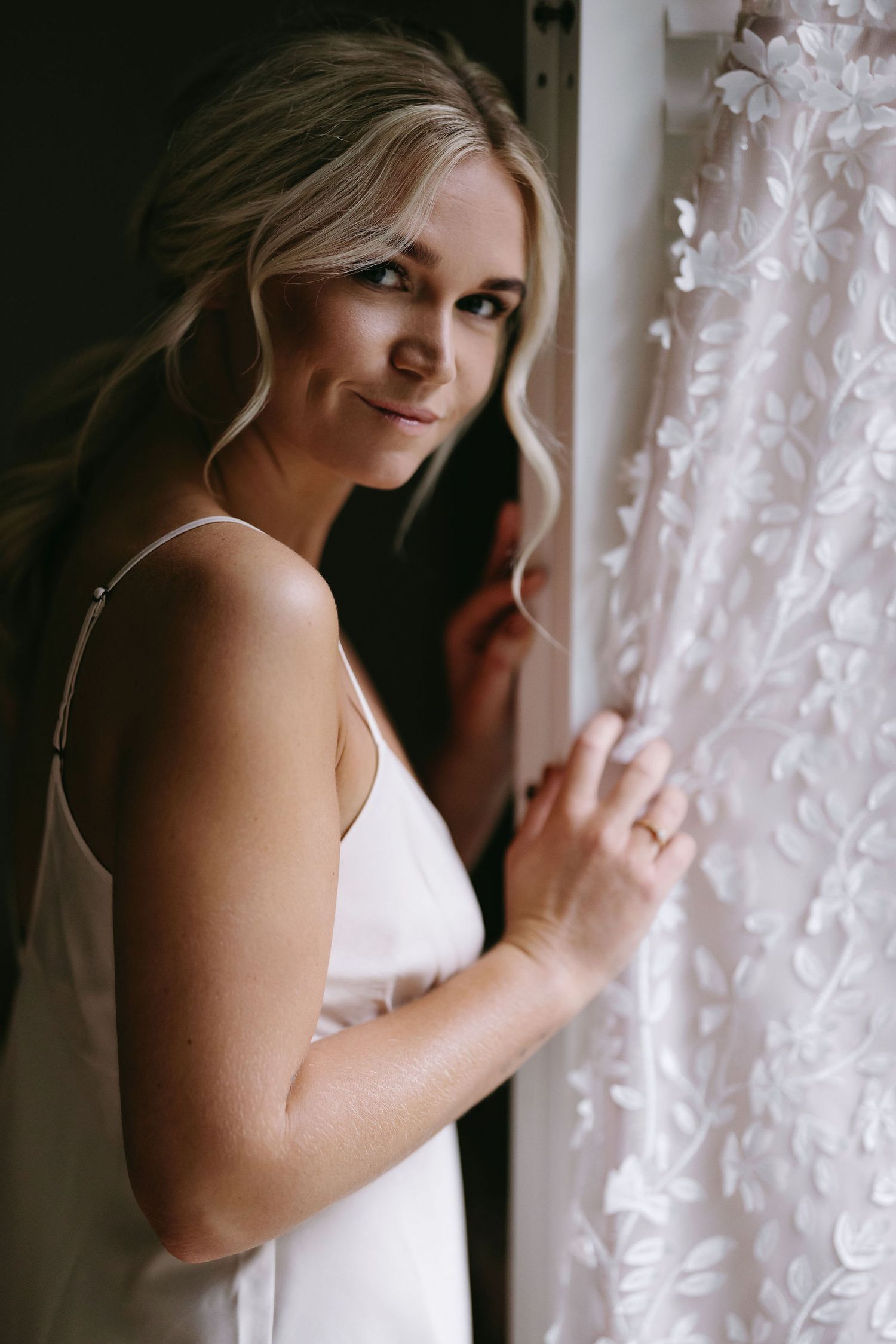 Elegant bride in white dress poses near window with wedding gown.
