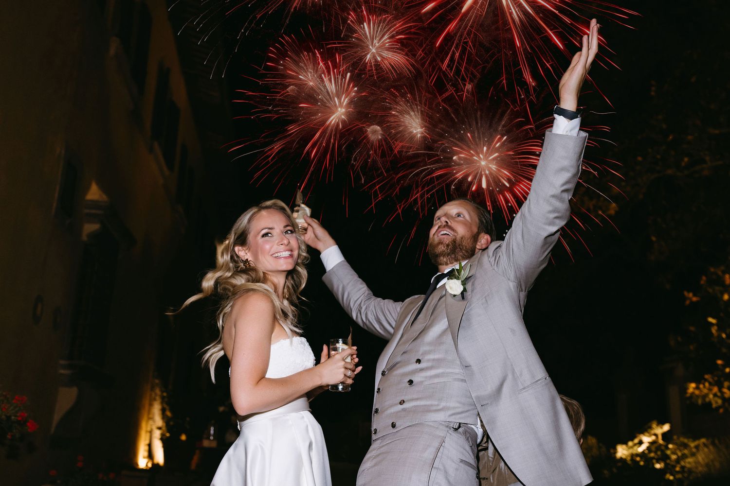 A couple celebrates during their wedding reception with fireworks bursting in the night sky behind them.