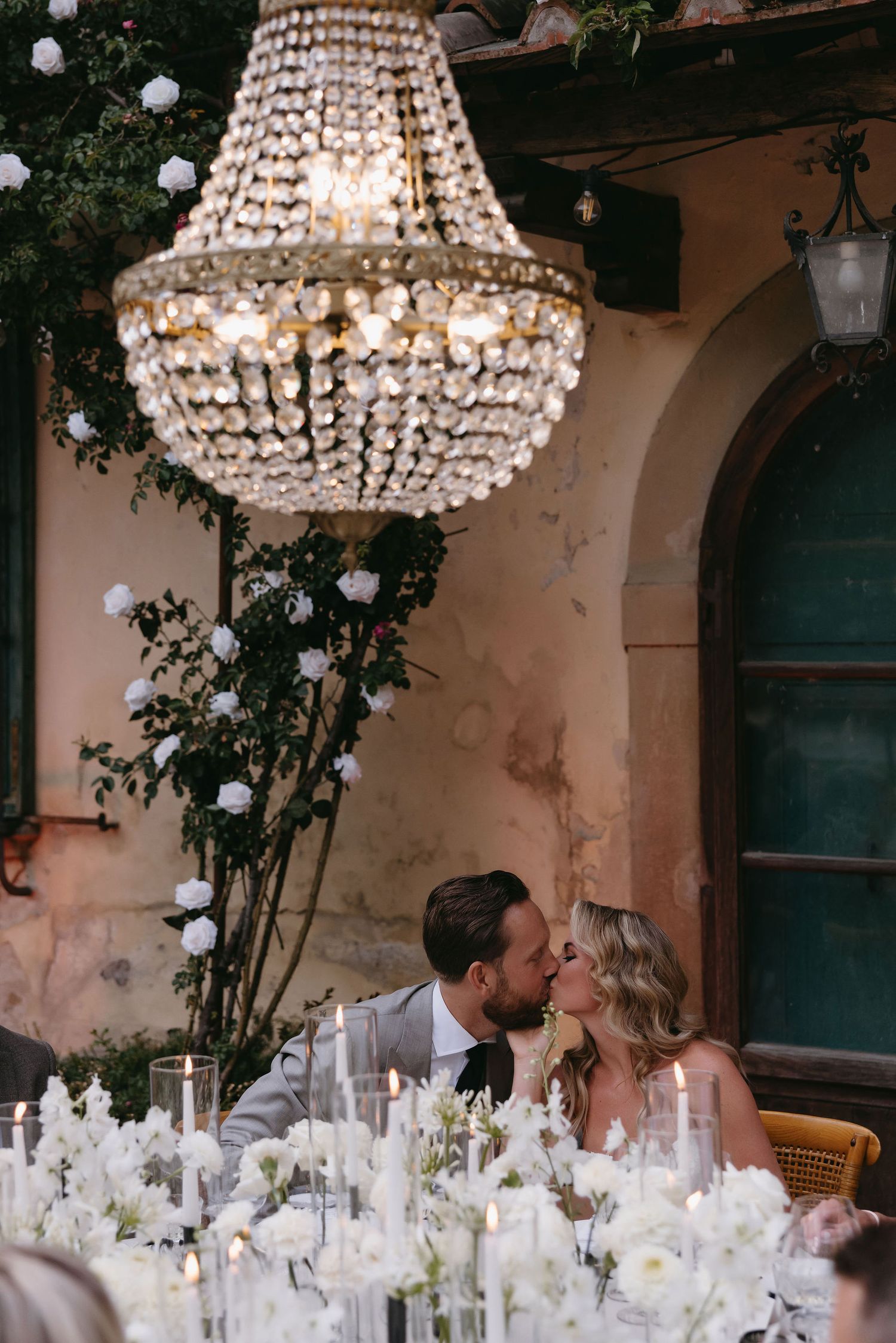 An elegant outdoor wedding reception under a crystal chandelier with white floral table decorations and peach-colored walls.