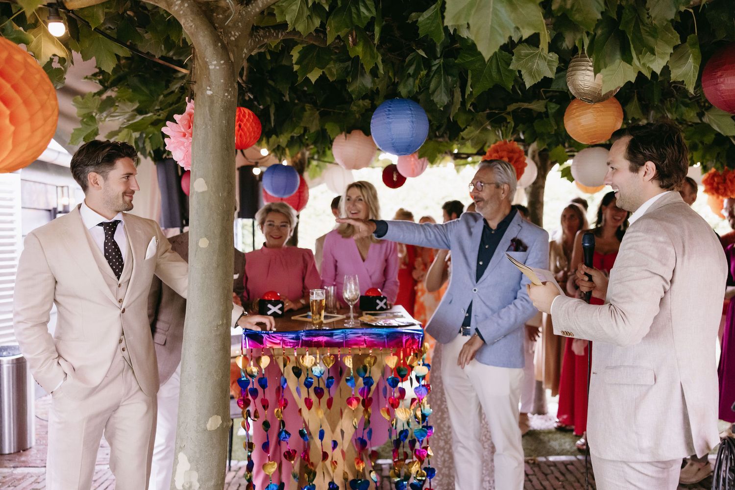 Outdoor wedding reception under trees decorated with colorful lanterns and string lights.