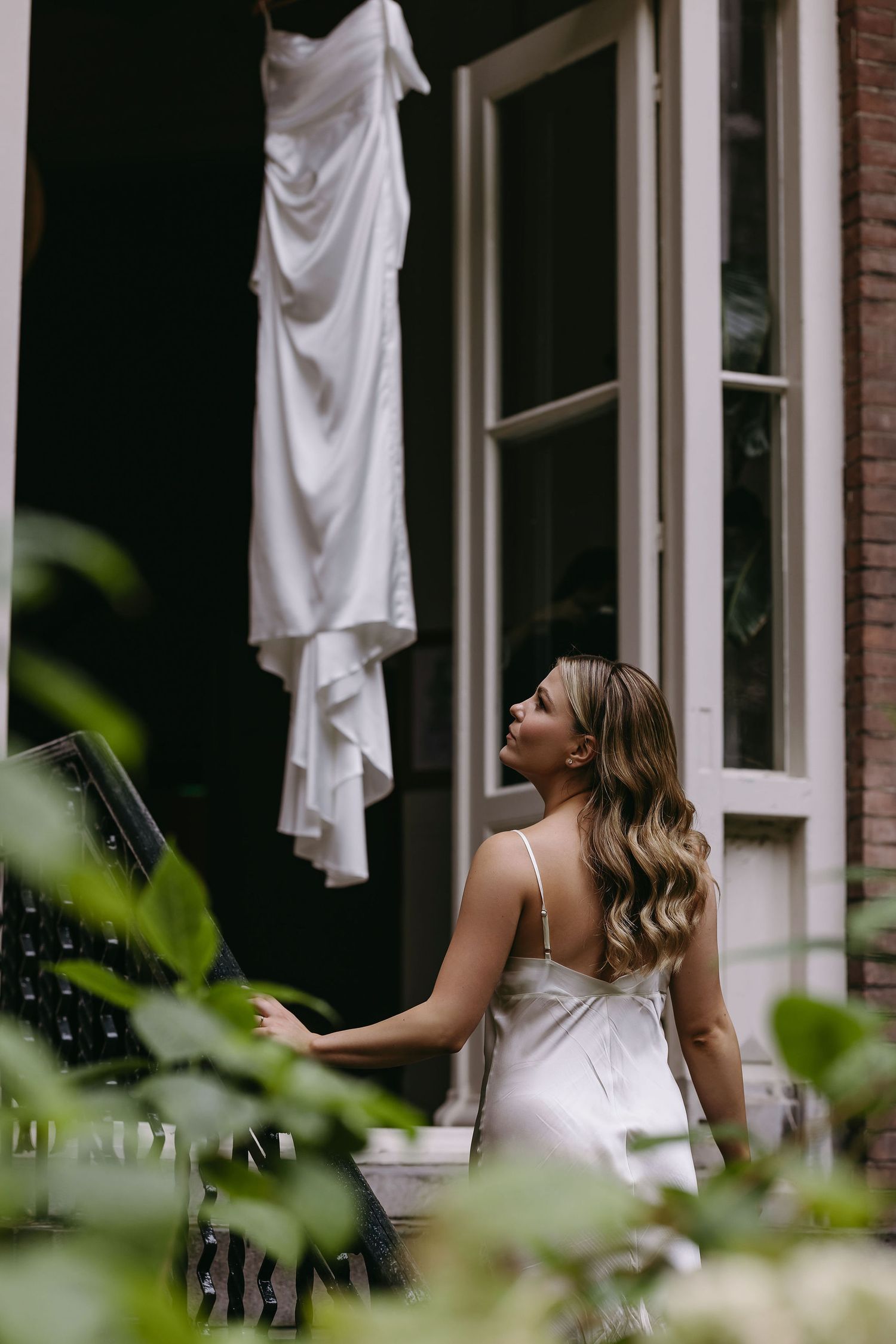 A white wedding dress hangs elegantly by a window while a figure in white admires it from below.