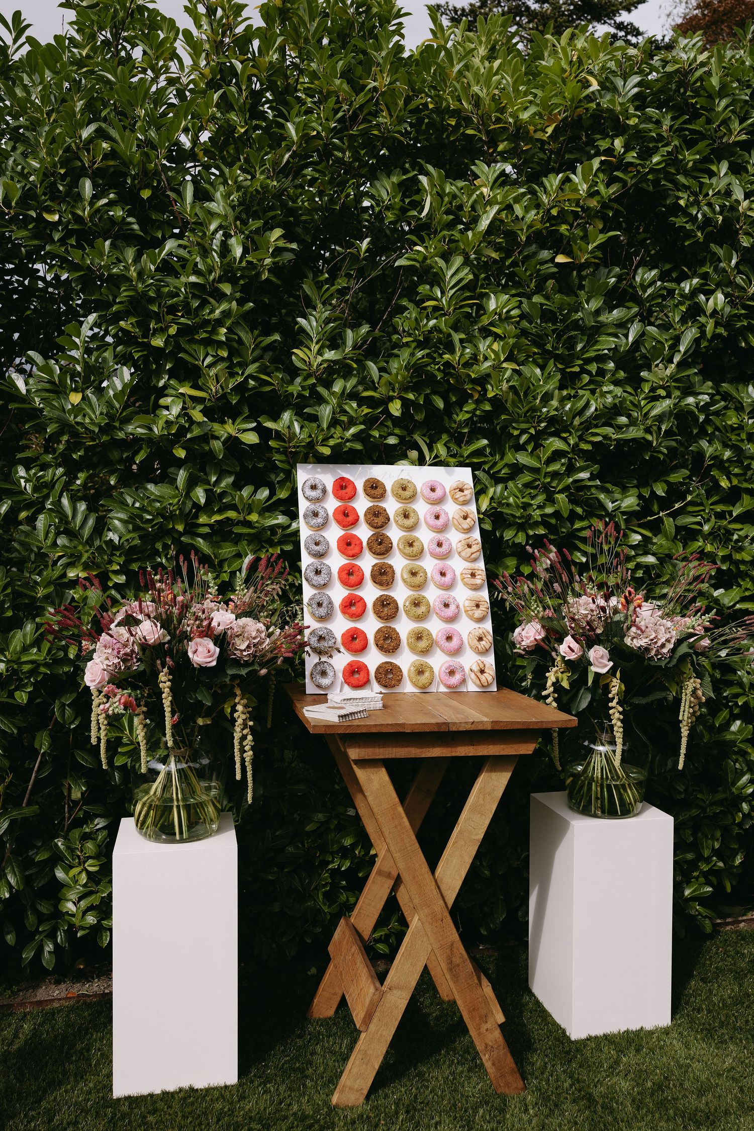 Wedding dessert display with donut wall and pink flower arrangements on white pedestals against green hedges.