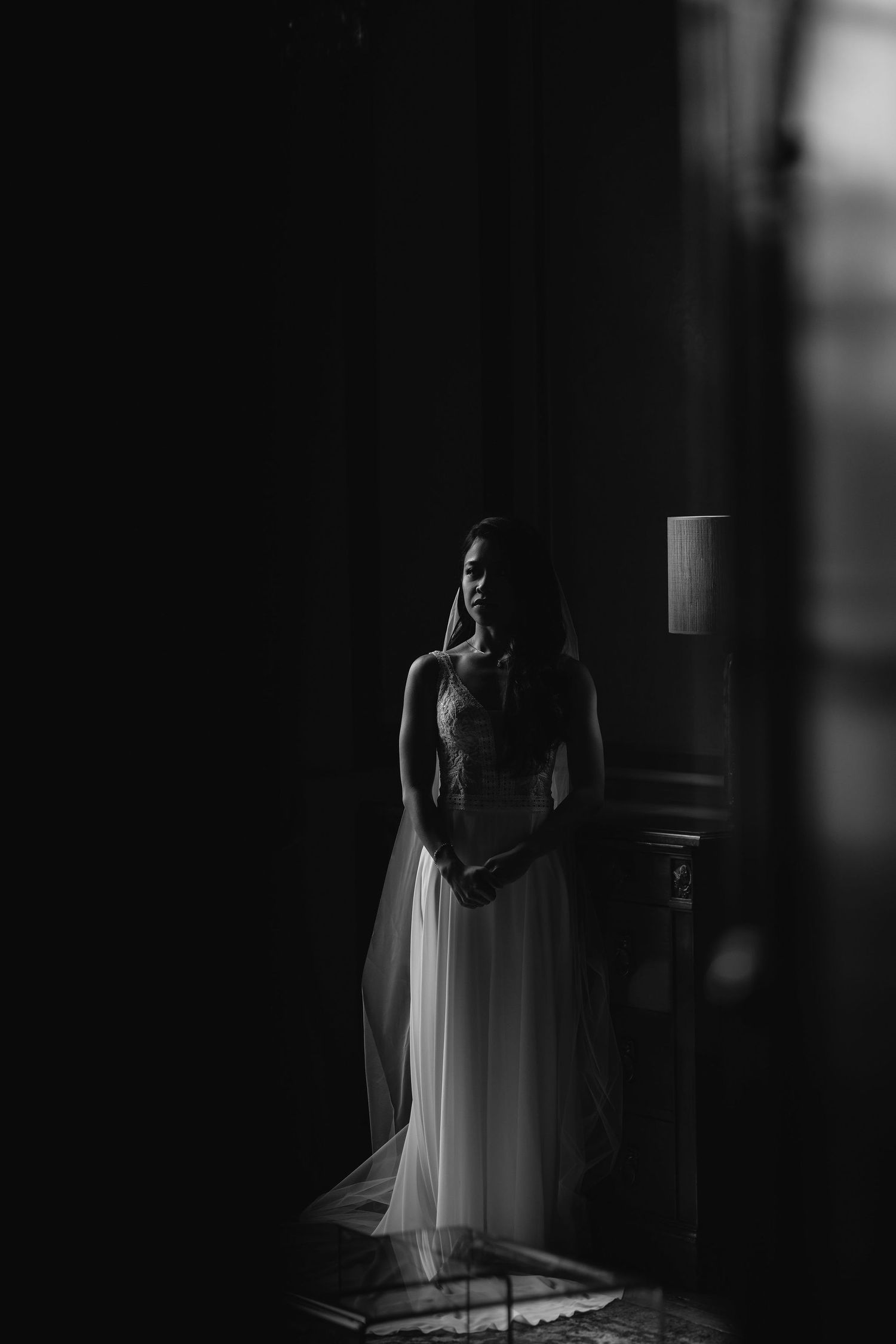 A person stands in a shadowy doorway wearing an elegant white dress in an artistic black and white composition.