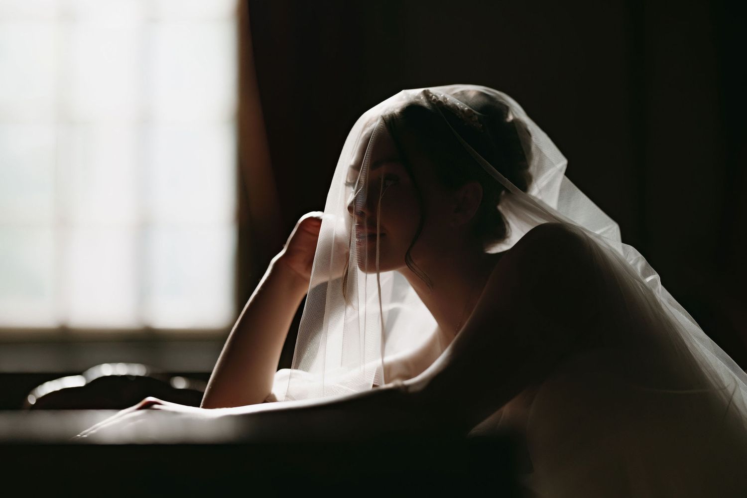 A silhouetted figure wearing a bridal veil sits in moody lighting by a window creating a dramatic atmospheric scene.