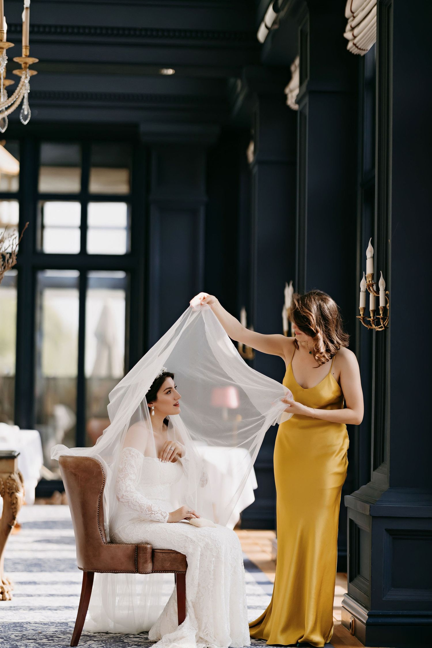 A bride in a white gown sits while an attendant in a golden dress adjusts her flowing veil in an elegant room.