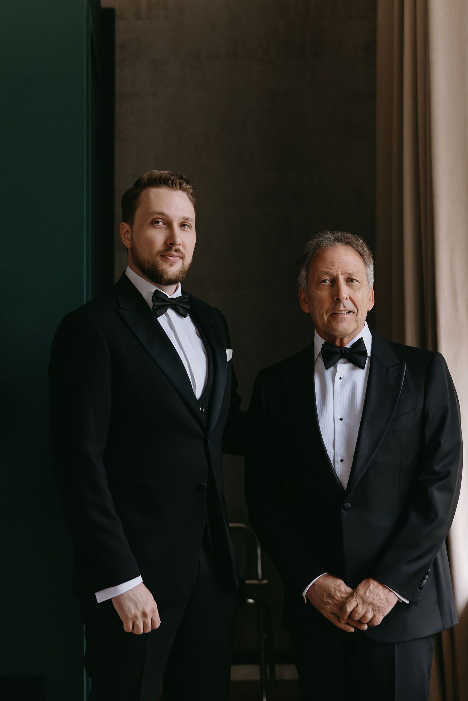 Two people in formal black tuxedos pose for elegant wedding portraits in a dimly lit setting.
