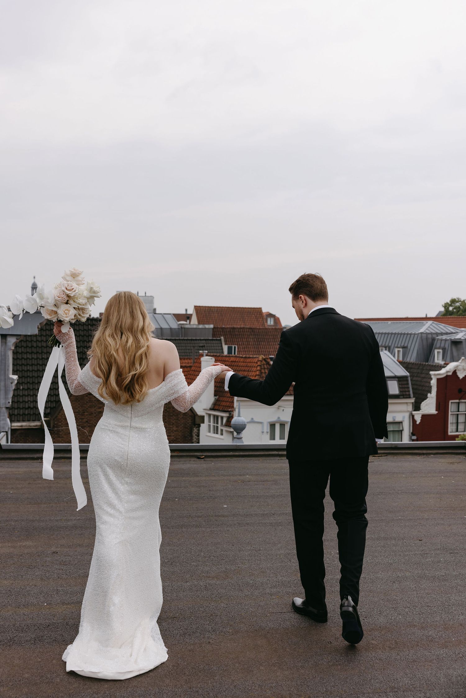 Couple in wedding attire walking together on a rooftop overlooking city buildings.