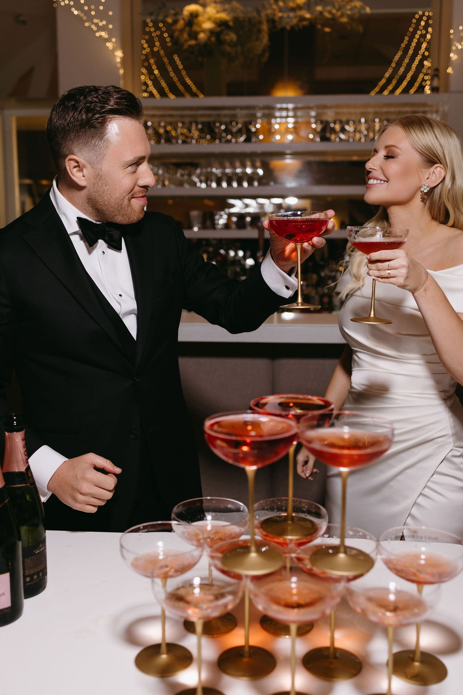 Couple toasts with pink cocktails at elegant wedding reception bar.