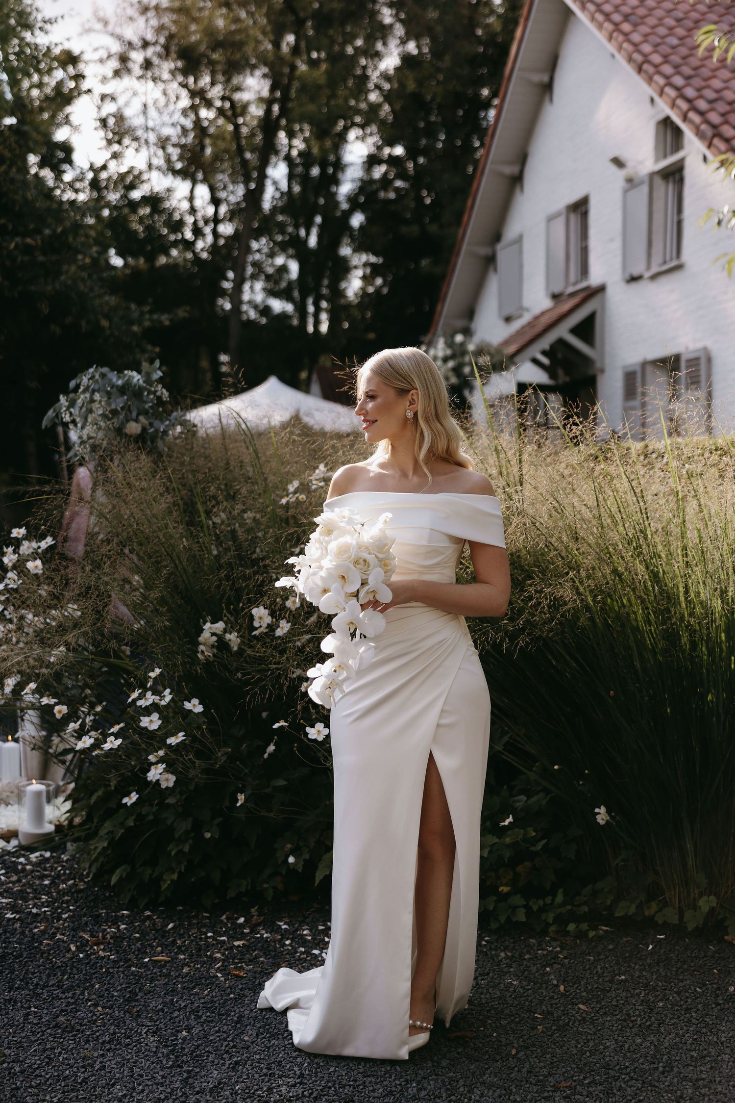 Elegant bride in white dress posing near ornamental grasses outside a white house.