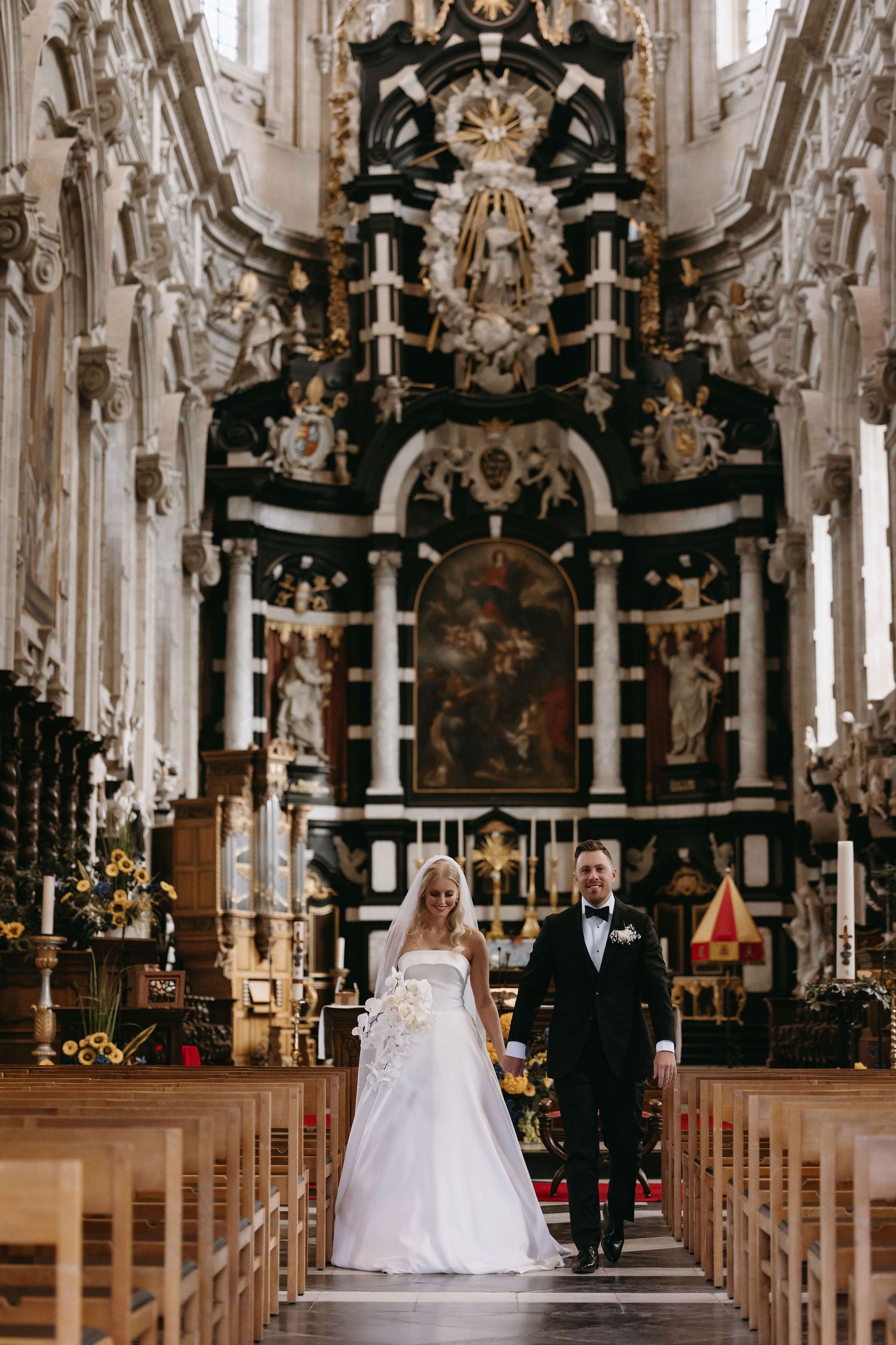 Couple walking down aisle in ornate baroque church with elaborate gold altar and decor.
