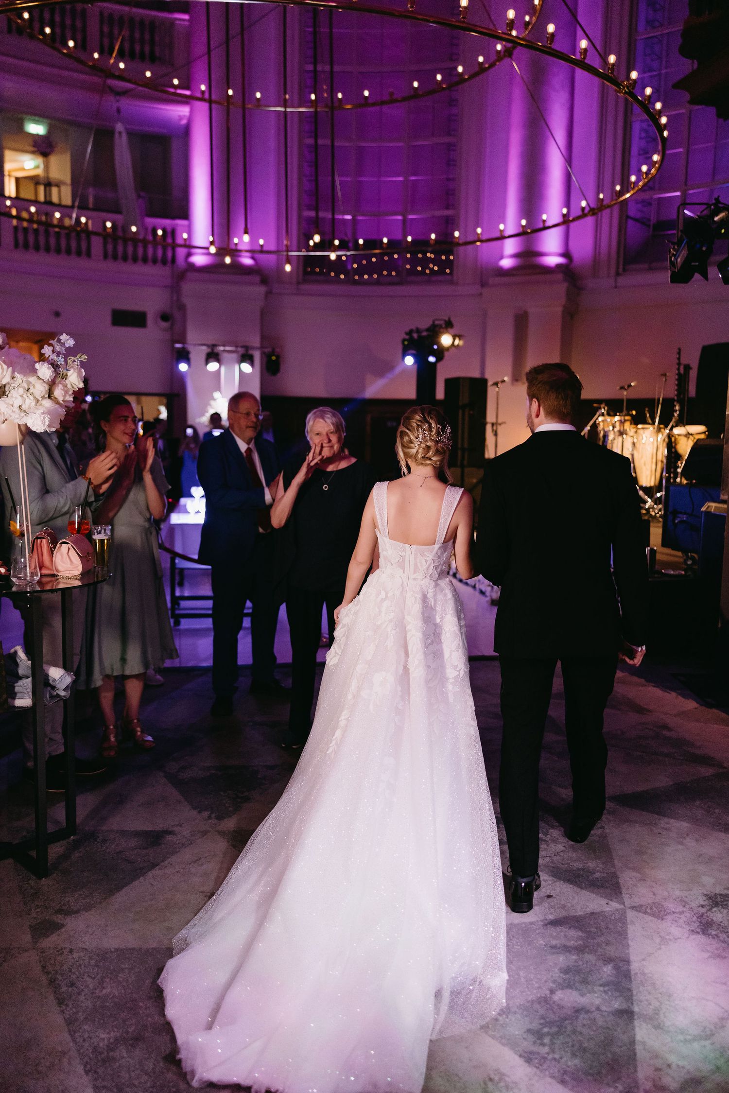 A bride in flowing white gown walks under purple lighting and string lights at reception venue.