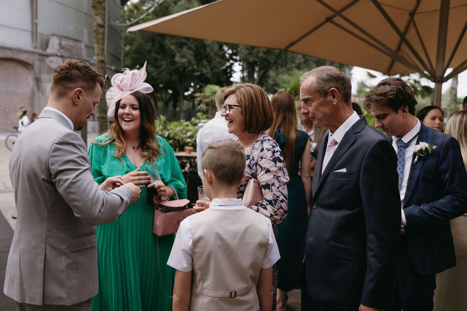 Wedding guests mingle during an outdoor cocktail reception under beige umbrellas.